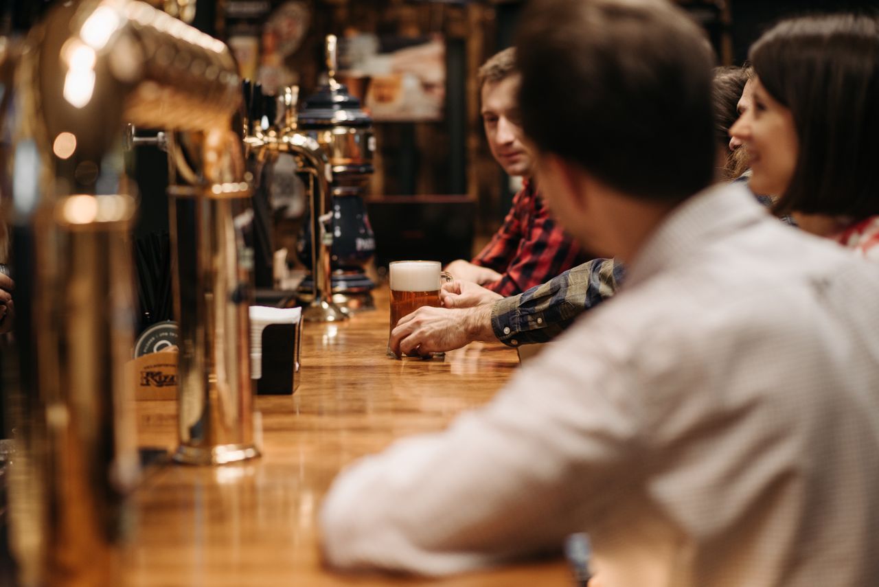 services-01 Friends enjoying a beer at a bustling bar with wooden counter and brass taps.