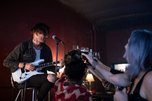 Musician playing guitar in a lively bar as people raise their glasses in celebration.